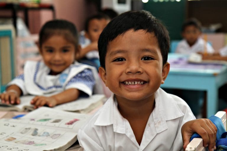 En pojke som ler och tittar in i kameran. I bakgrunden sitter en flicka. De befinner sig i ett klassrum.