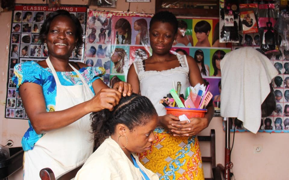 Tre kvinnor i en frisörsalong. Ena kvinnan ler medan hon flätar en av de andra kvinnornas hår.