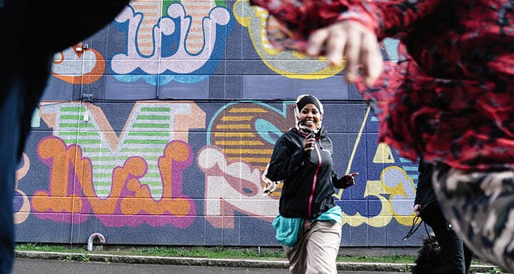 En glad kvinna springer mot kameran. En graffitimålad vägg syns i bakgrunden.