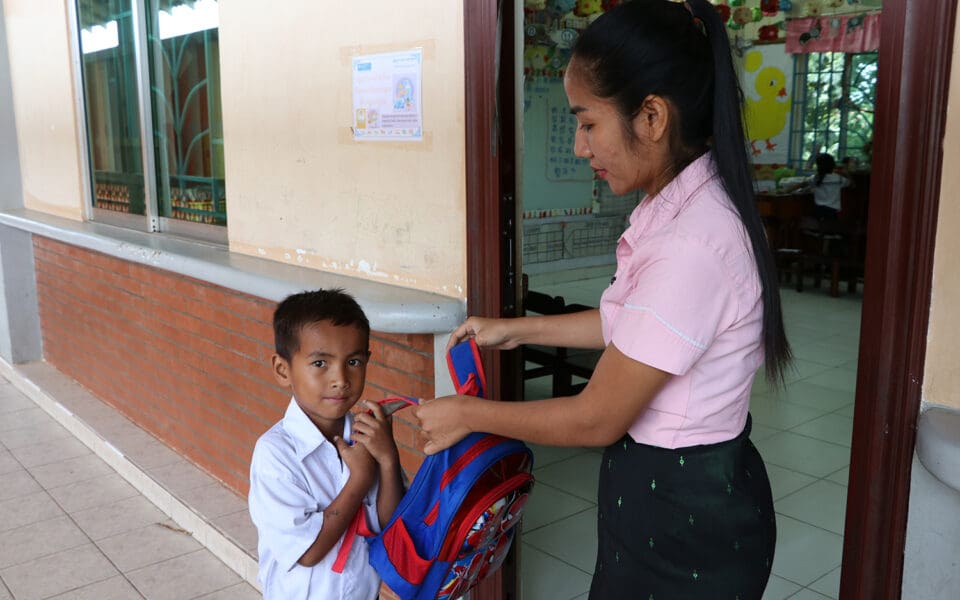 En lärare hjälper en kambodjansk pojke att ta på sig ryggsäcken.