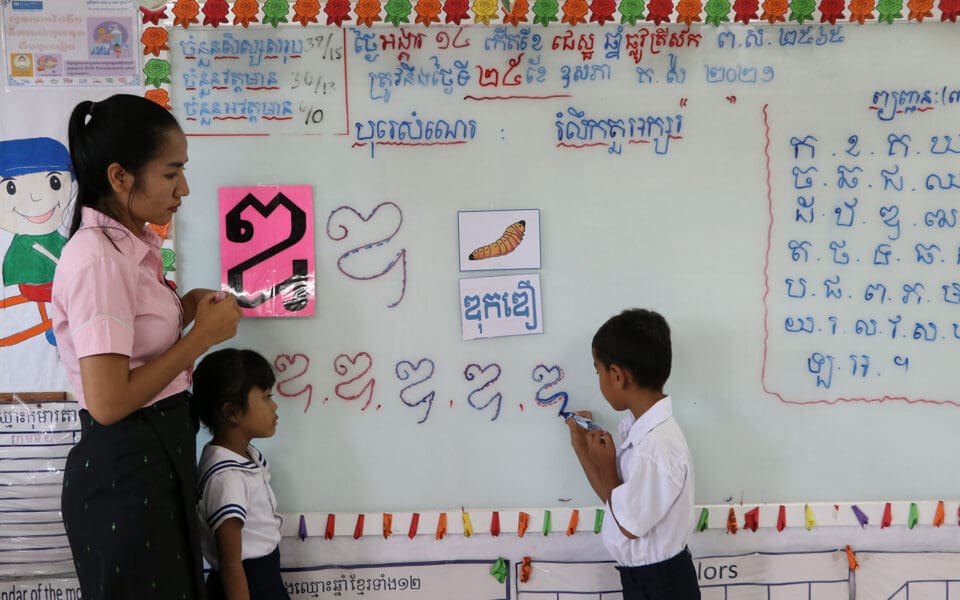två kambodjanska barn står vid en skoltavla. Läraren står till vänster om dem.