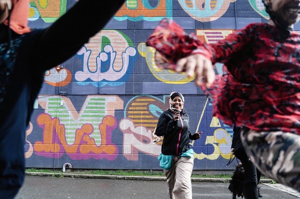 Tre personer som springer mot kameran. I bakgrunden syns en husfasad med graffiti.