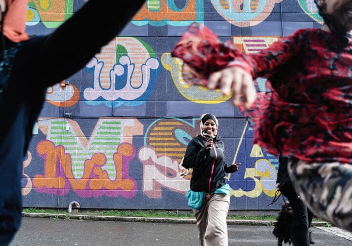 Tre personer som springer mot kameran. I bakgrunden syns en husfasad med graffiti.