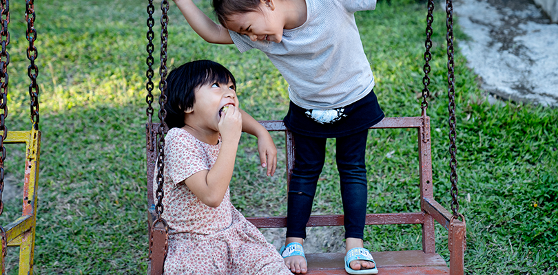 Två små barn skrattar mot varandra och står och sitter på en gunga med grönt gräs i bakgrunden