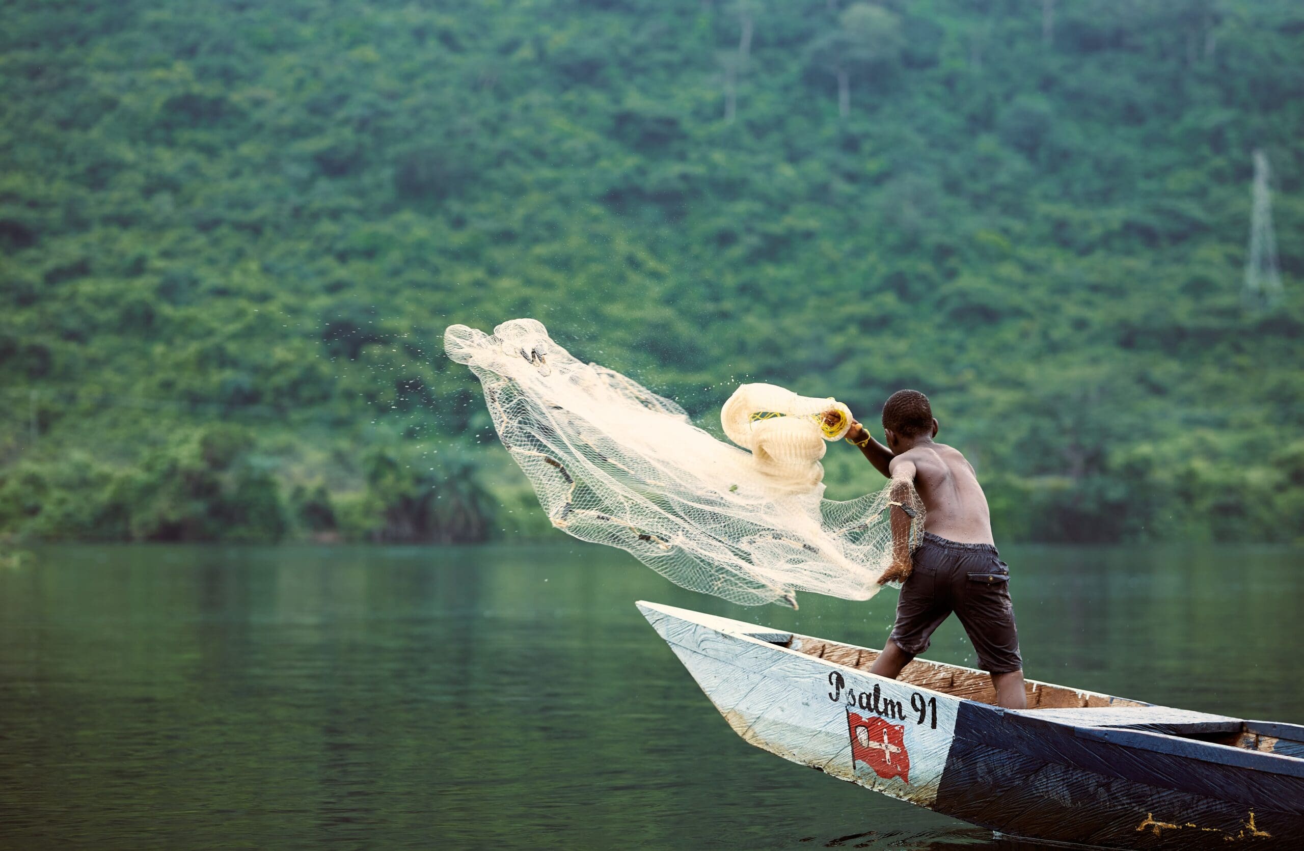 Pojke i bar överkropp i en ranglig båt kastar ut ett fiskenät i en mörk flod