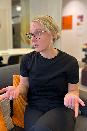 Kvinna i ljust hår och svart tröja pratar och gestikulerar i en soffa