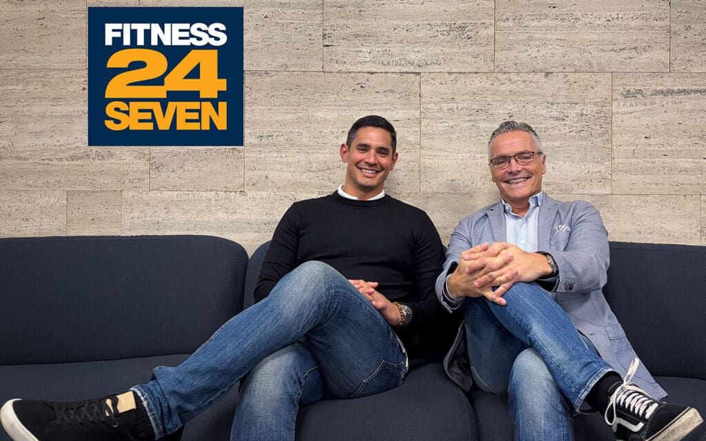 Två män ler i soffa med grå vägg i bakgrunden